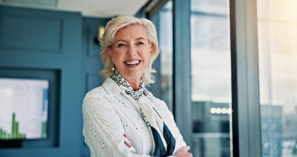 A portrait of a smiling senior woman with blonde hair, wearing a white patterned shirt and a scarf, standing in a modern office with her arms crossed.