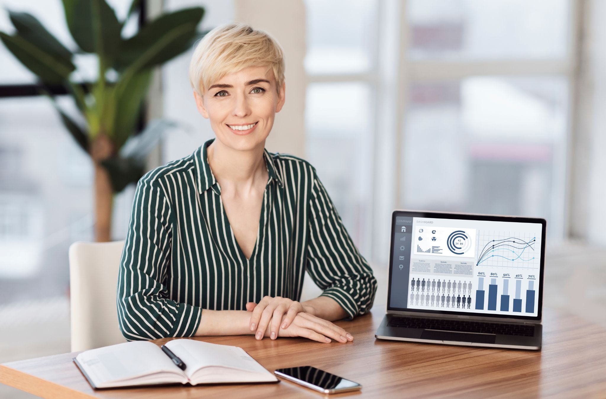 A smiling woman with short blonde hair sitting at a table with an open laptop displaying data charts.