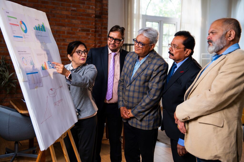 A woman pointing at charts on a presentation board during a business meeting with four male colleagues looking on.