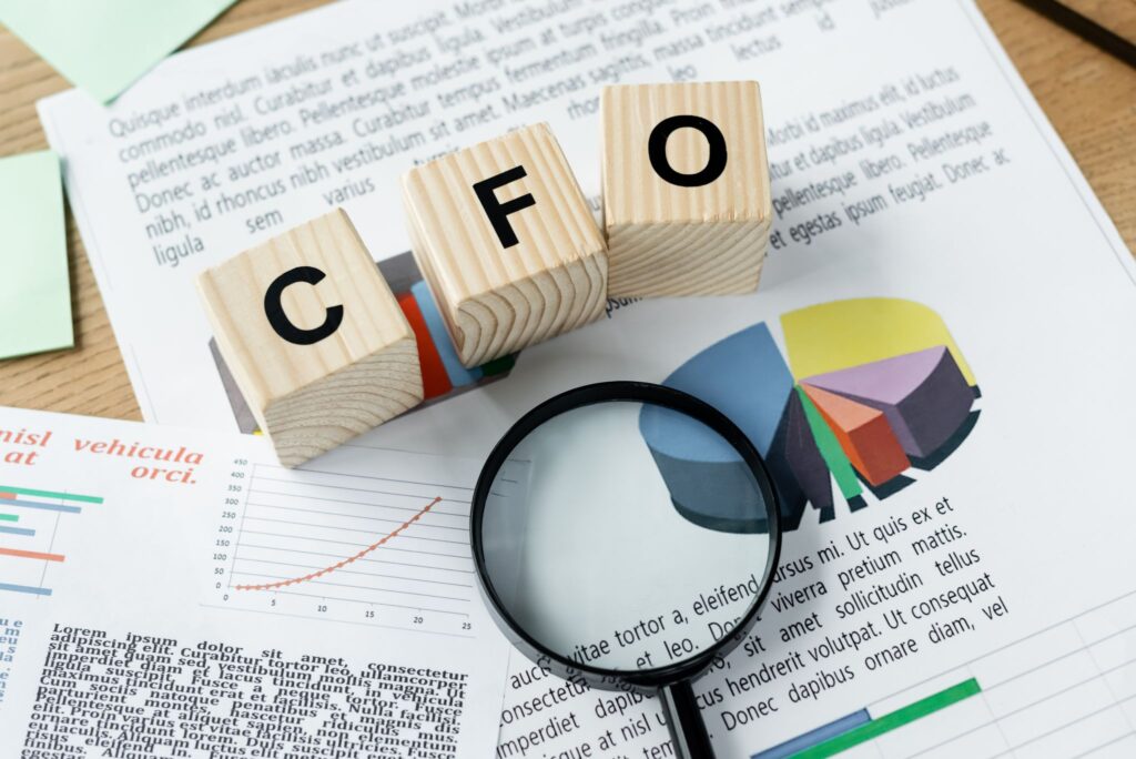 Three wooden blocks spelling out CFO resting on top of financial documents with pie charts and graphs, with a magnifying glass placed in the foreground.