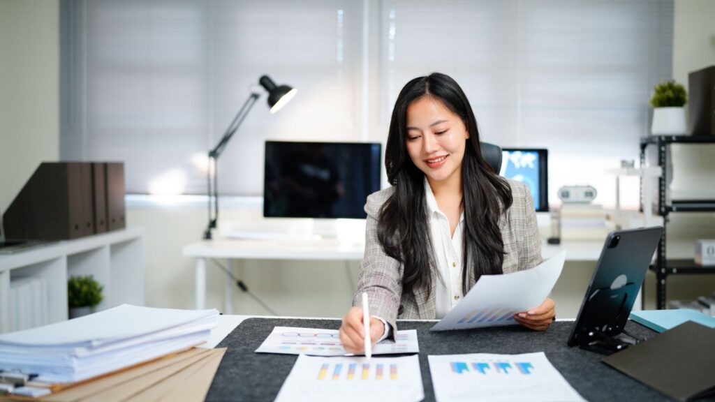 An Asian businesswoman reviewing financial performance charts and working on documents in a modern home office.