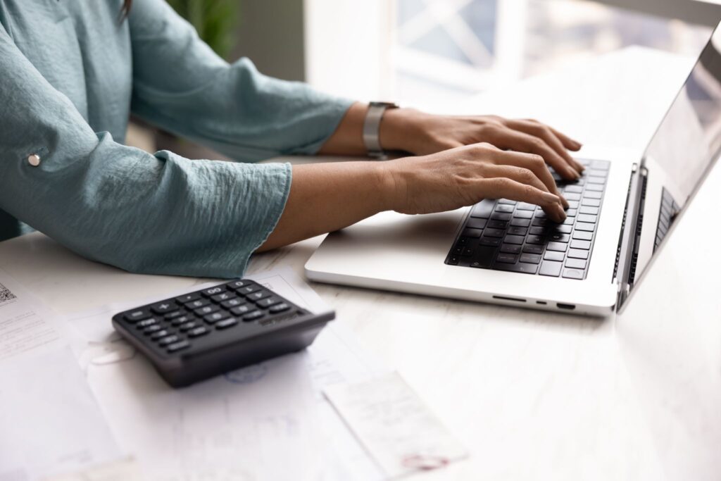 Close-up of a professional typing on a laptop at a desk with a calculator and financial papers nearby.