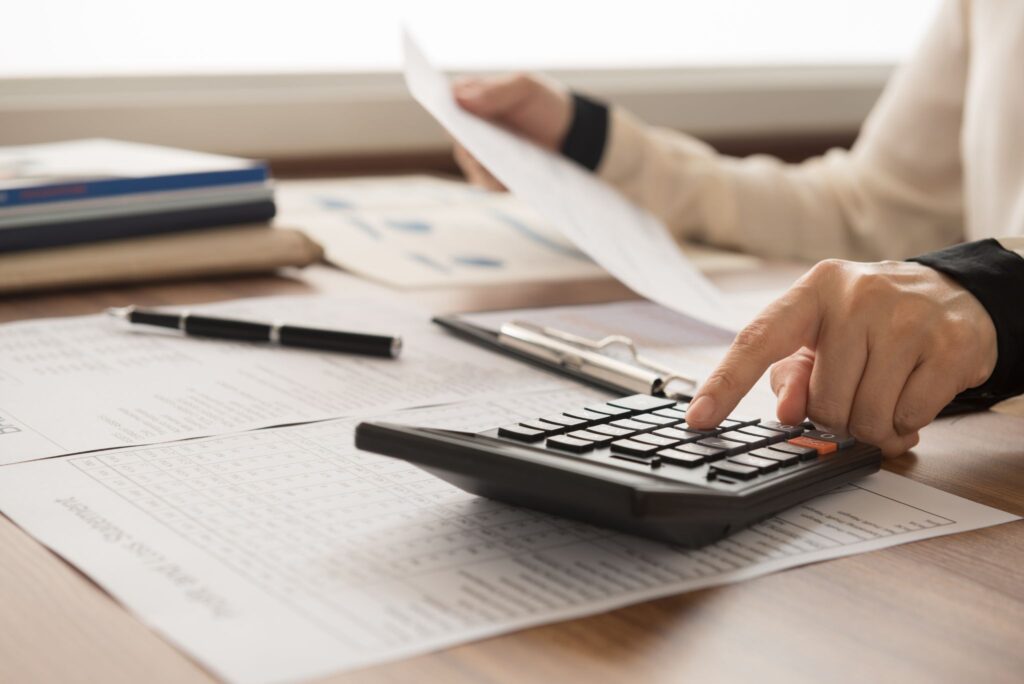 Close-up of hands using a desktop calculator while reviewing financial documents and spreadsheets at a desk.
