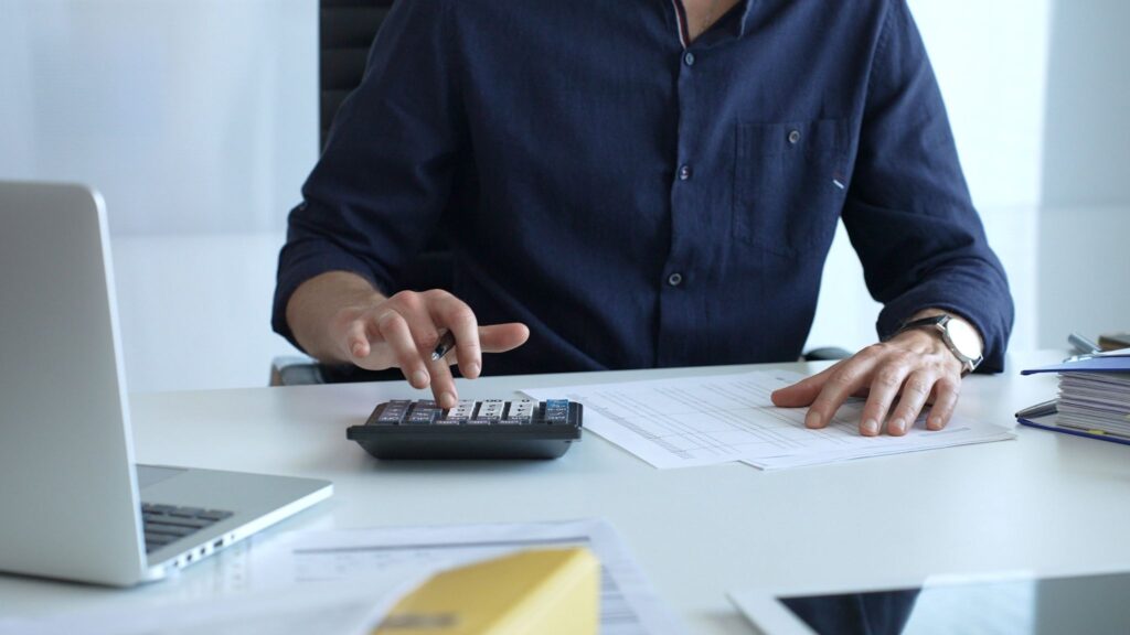Man sitting at a desk using a calculator while reviewing financial documents and spreadsheets.