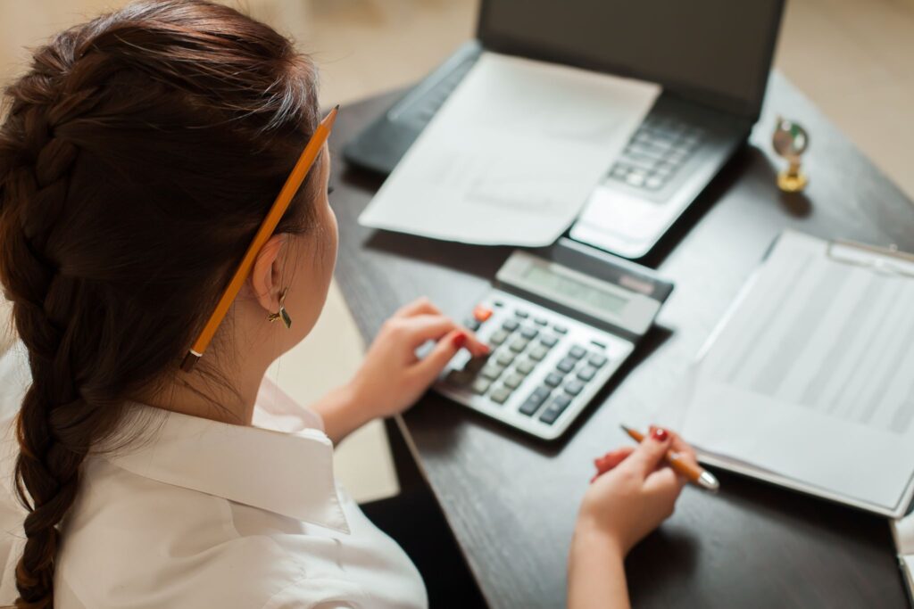 Over-the-shoulder view of a female accountant using a calculator at a desk with a laptop and financial documents.