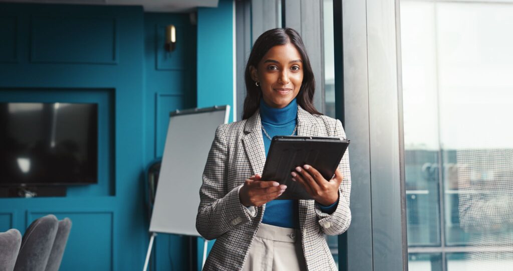 Professional woman standing near a window in a modern office holding a digital tablet.