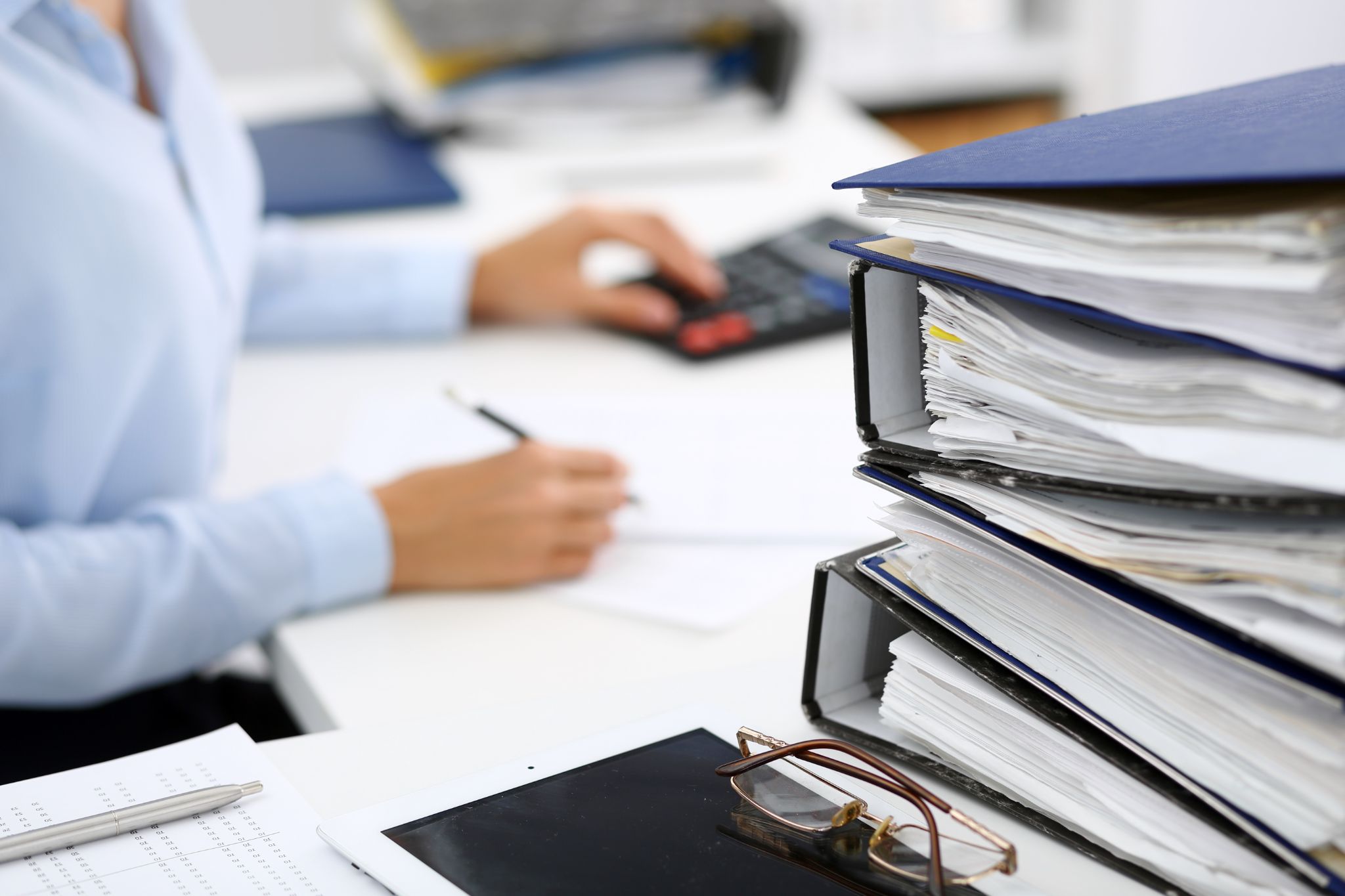 Stack of accounting binders on a desk with a professional working on tax or financial paperwork in the background.