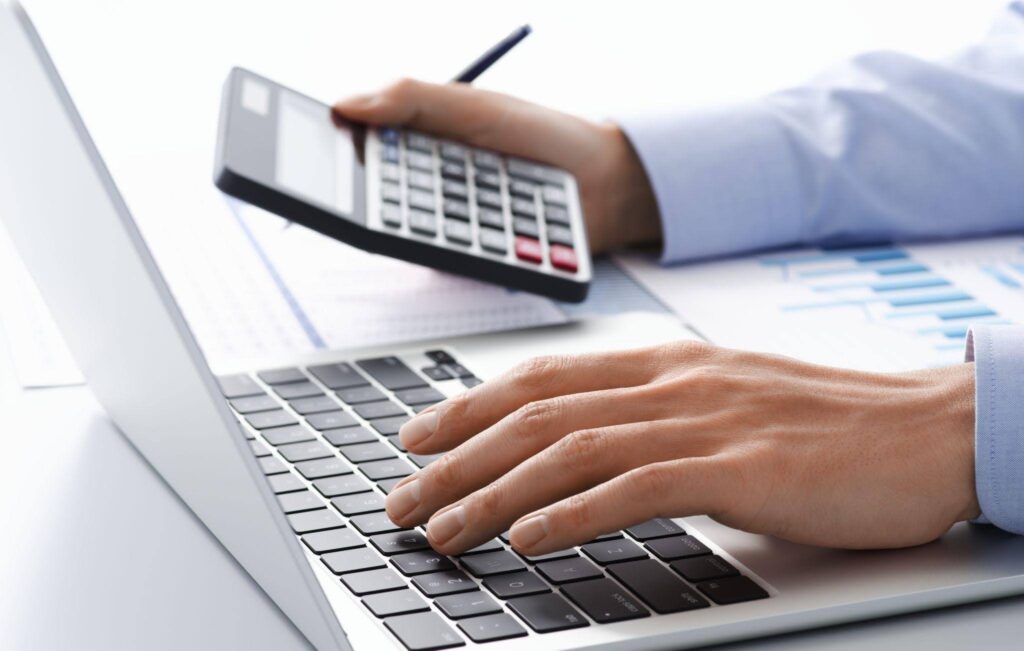 A person typing on a laptop while using a calculator on the desk focused on their work