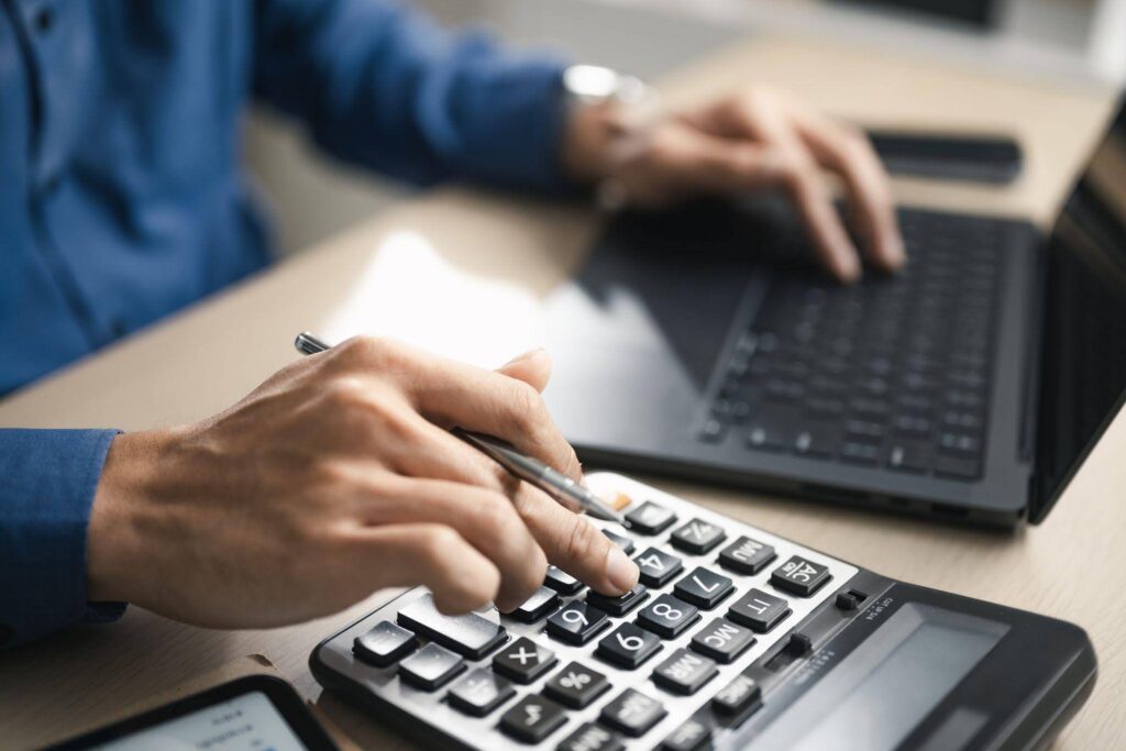 A person using a calculator while working on a laptop focused on their calculations and tasks