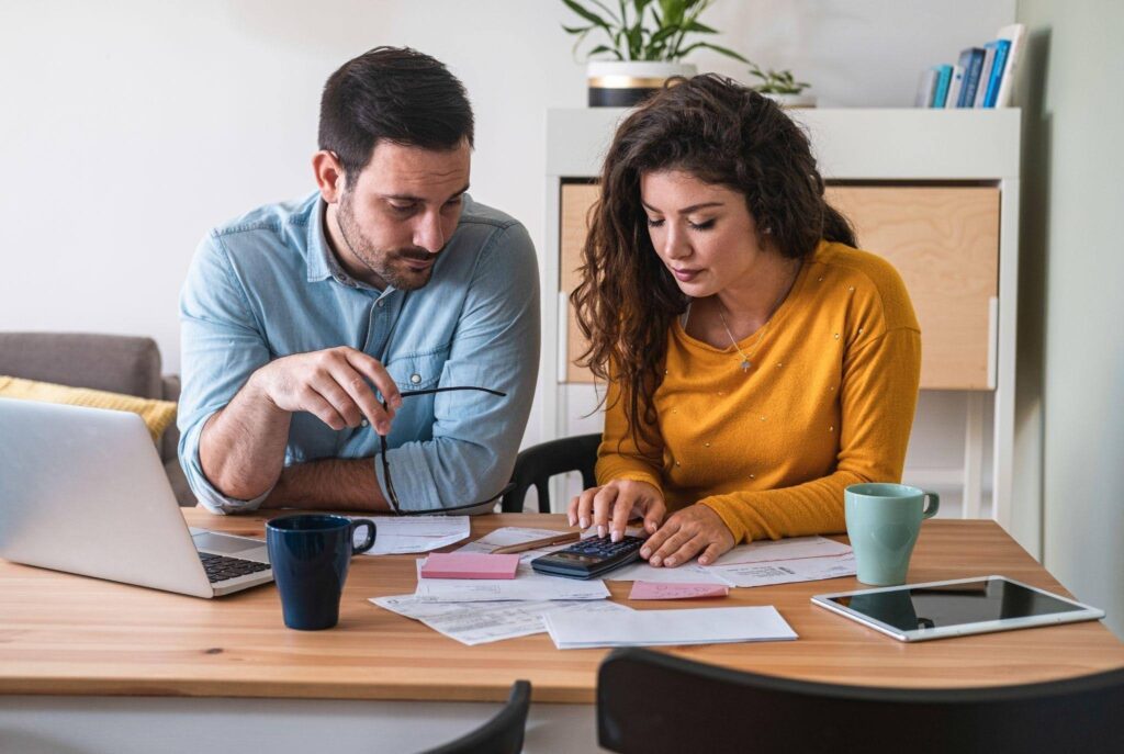 A man and woman sit at a table focused on a laptop engaged in discussion