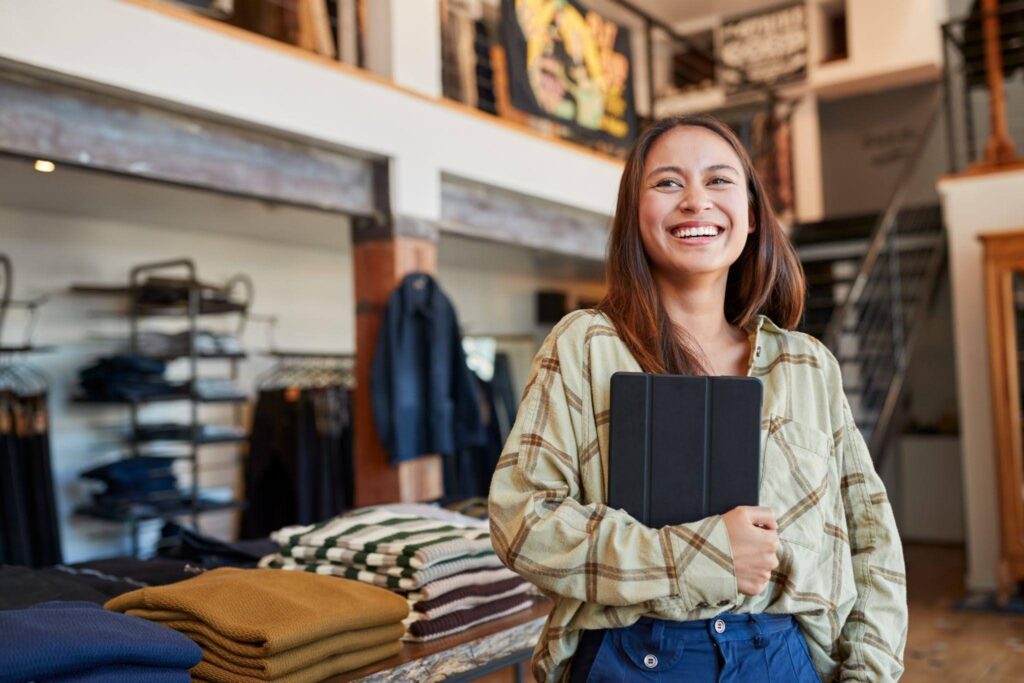 A woman stands in a clothing store holding a tablet and browsing through fashion options