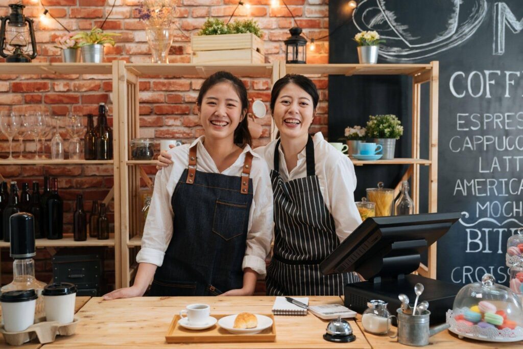 Two Asian women smiling at the camera standing in front of a coffee shop with outdoor seating