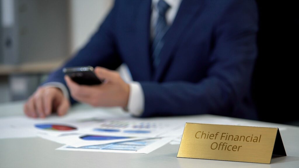 A close-up of a gold Chief Financial Officer desk nameplate, with a blurred person in a blue suit holding a smartphone in the background.
