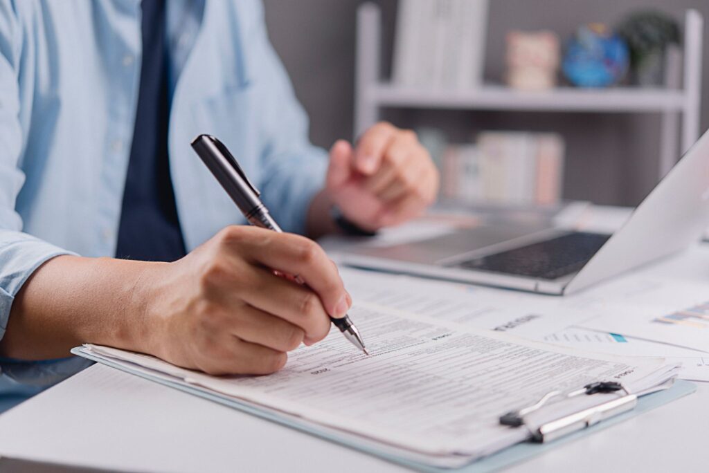 A close-up shot of a person's hand holding a pen and filling out a document on a clipboard, with a laptop open on the desk in the background.