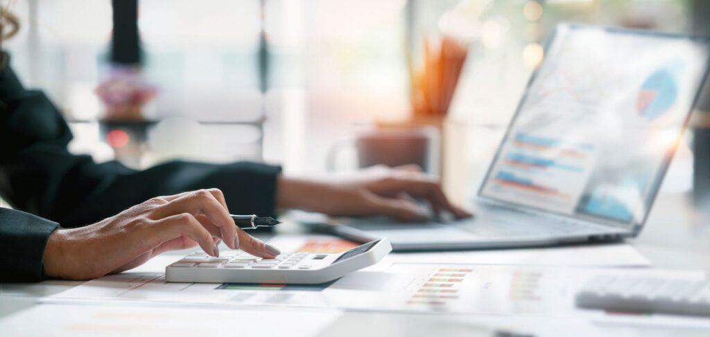 Accountant using a calculator while analyzing financial reports and working on a laptop.