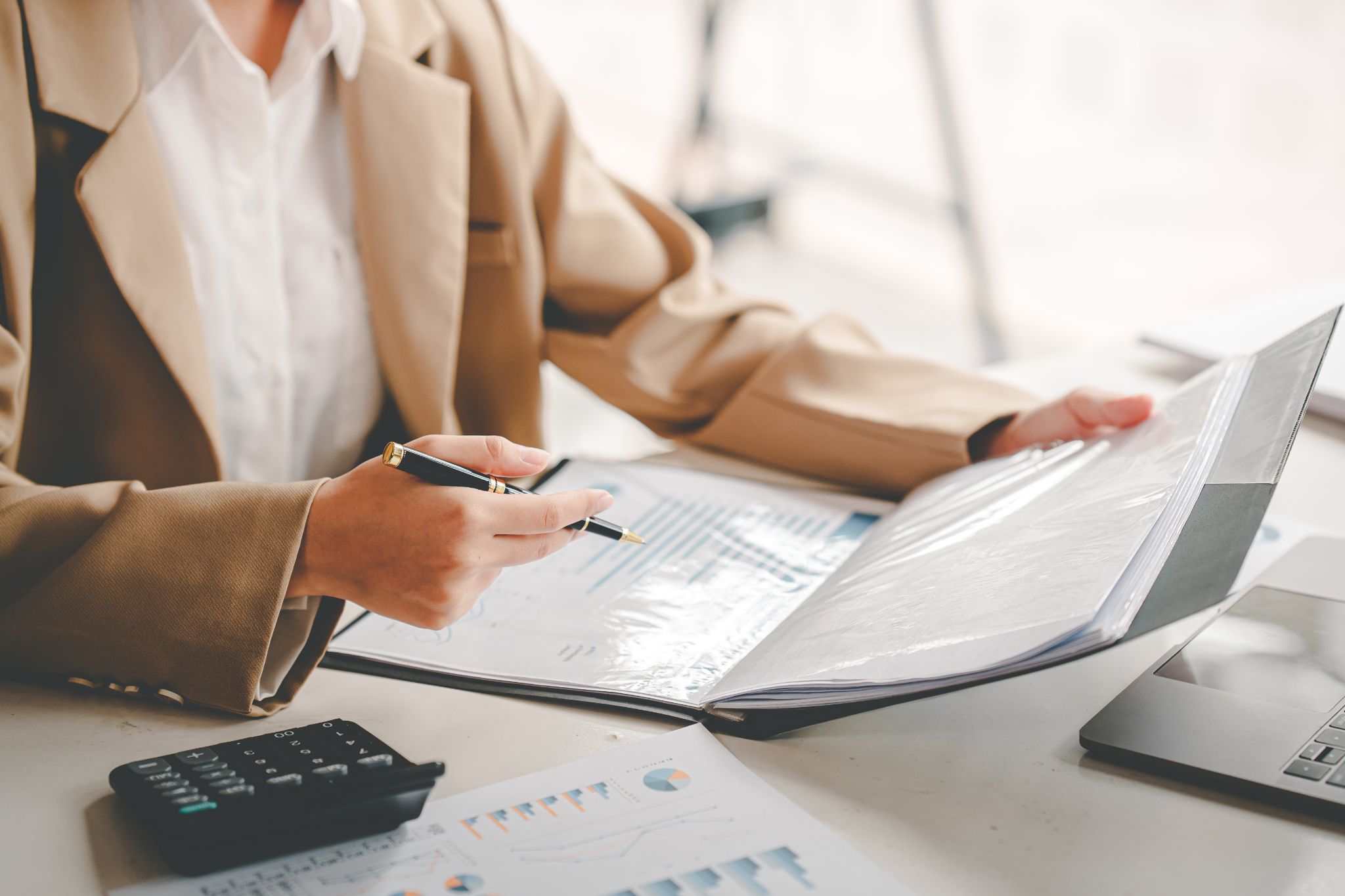 Business professional in a beige blazer holding a pen while reviewing financial reports in a binder at a desk with a calculator and laptop.