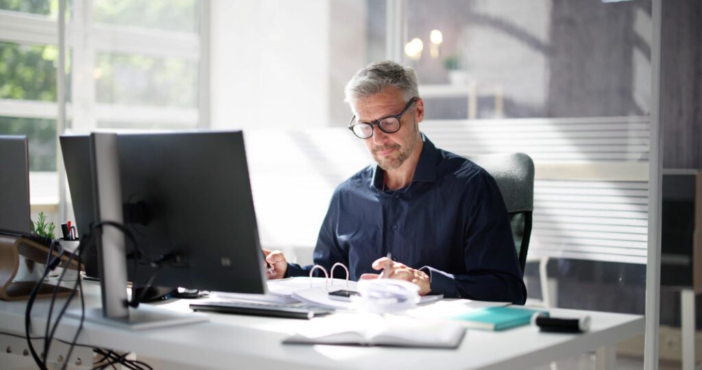 Mature businessman wearing glasses writing notes and reviewing paperwork at his office desk.