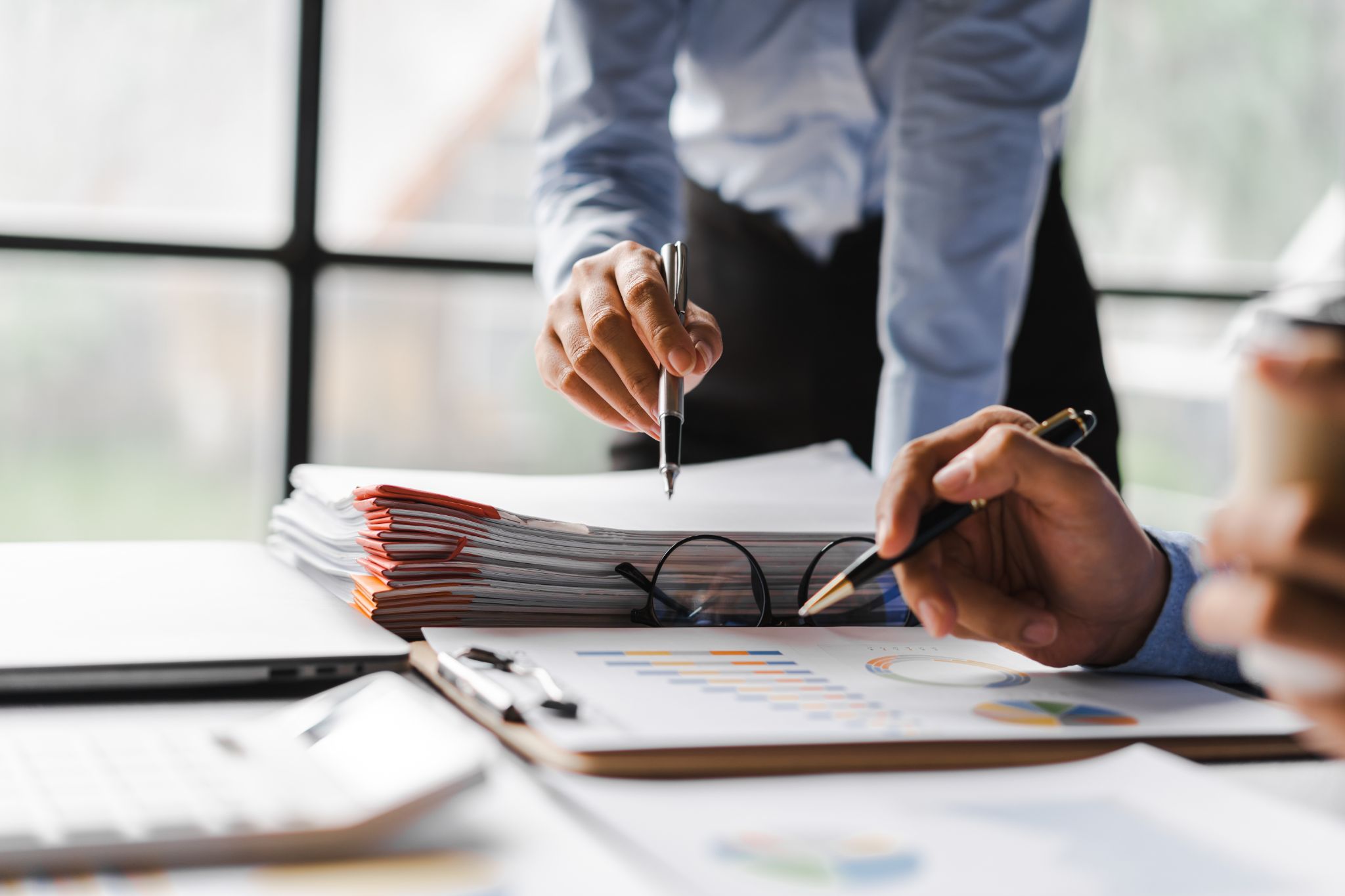 Two colleagues pointing with pens at financial charts and a stack of paperwork during a meeting.