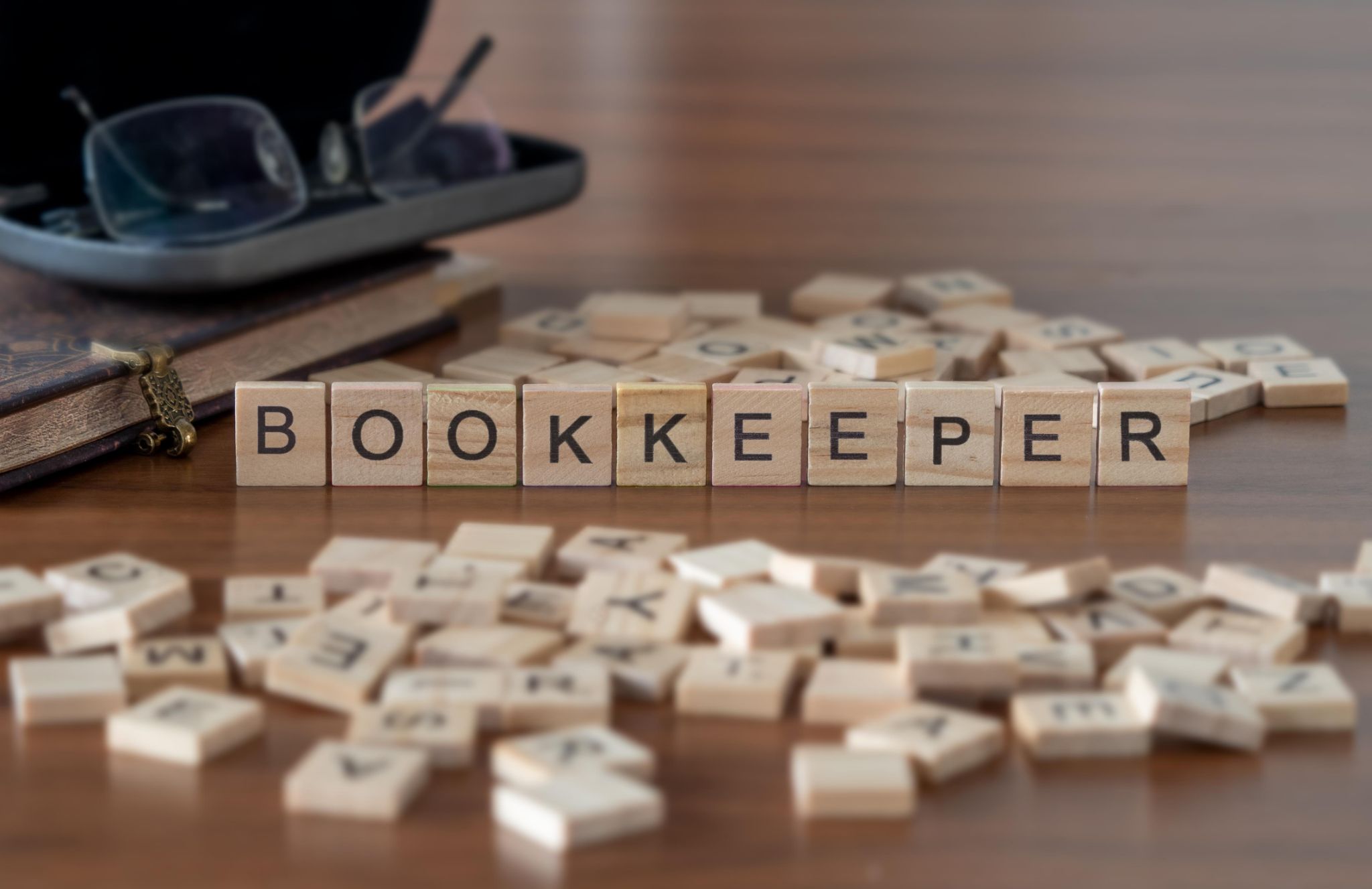 Wooden letter tiles spelling the word BOOKKEEPER arranged on a wooden table next to reading glasses and a notebook.